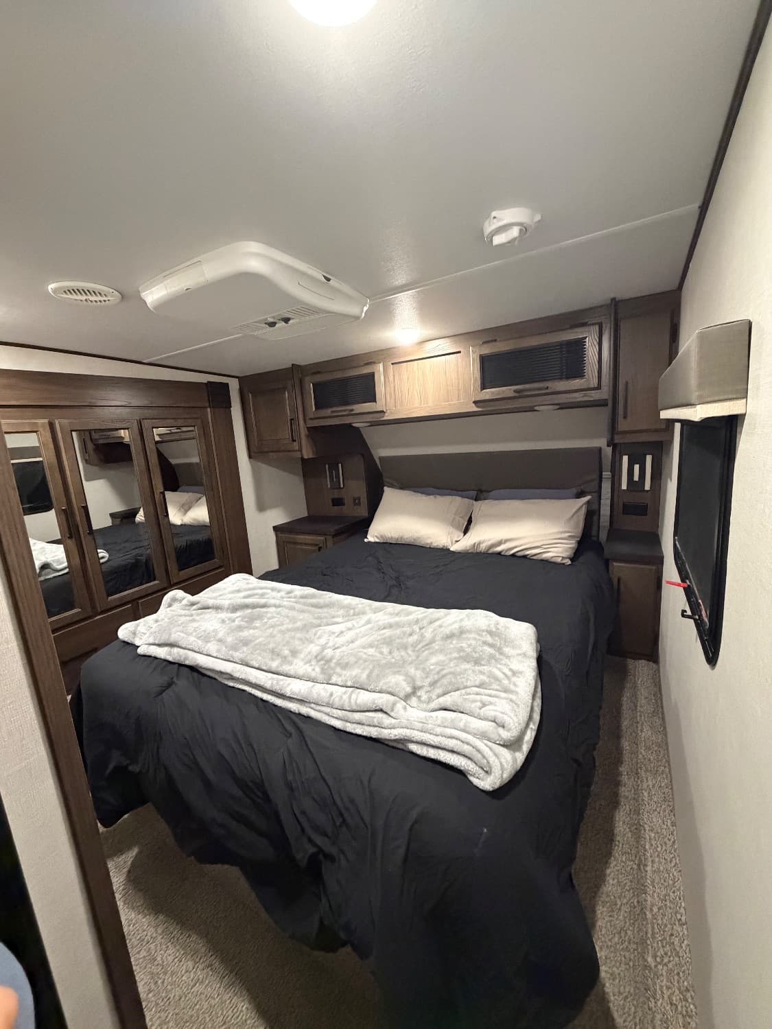 The queen master suite in The Crusader, made up with a headboard and reading lights, overhead cabinets and a window above it, a mirrored wardrobe along one wall, a wall-mounted TV opposite and its own roof air conditioner in the ceiling