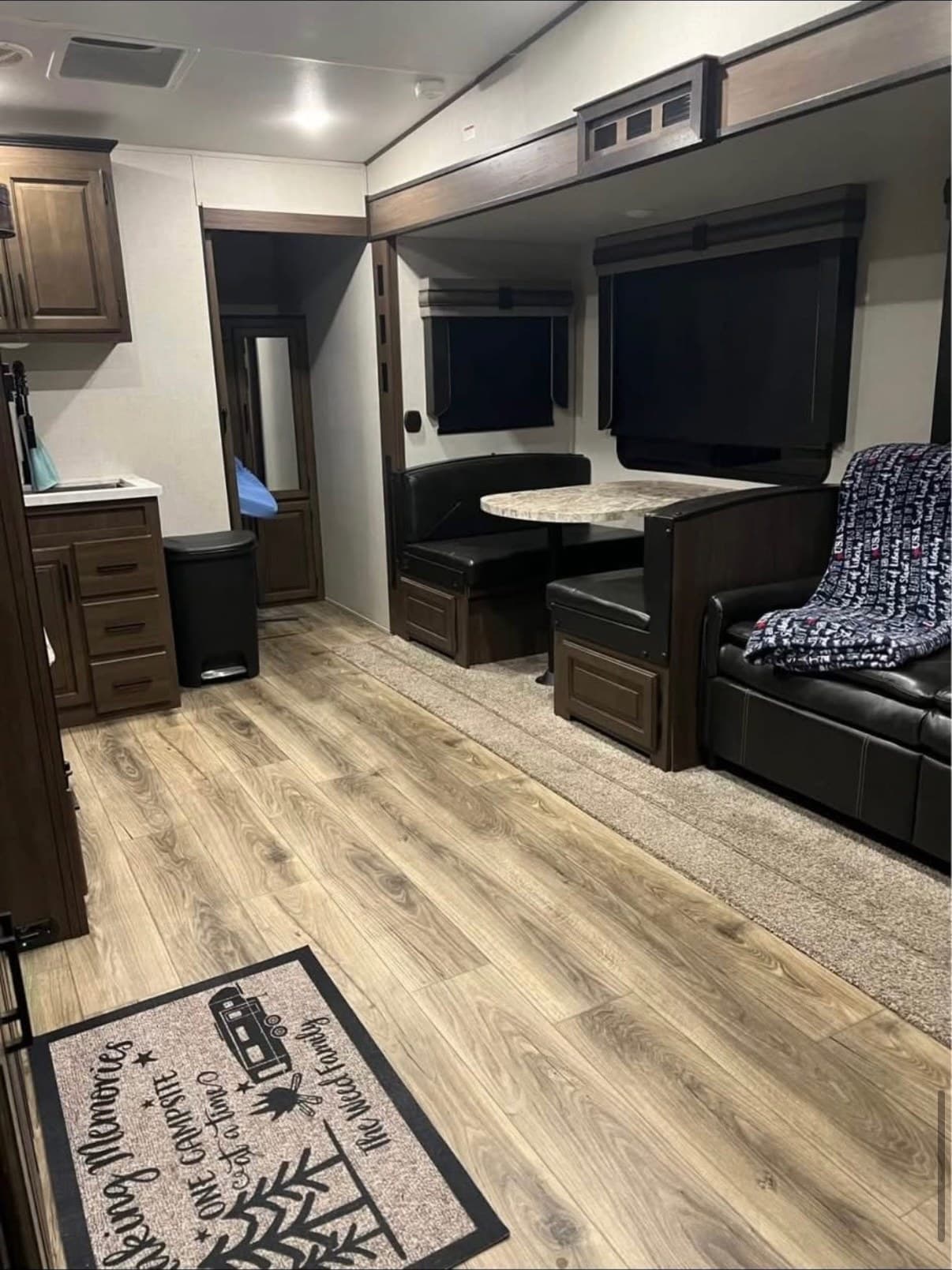 The living area of The Crusader with the full-size drop-down dinette and table in the slide-out, the queen sofa bed beside it, and wood-look flooring running through to the bathroom door